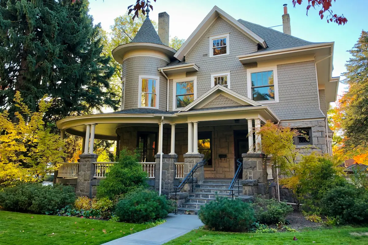 Victorian house with gray shingle siding, a round turret, white trim and a wraparound porch among autumn trees