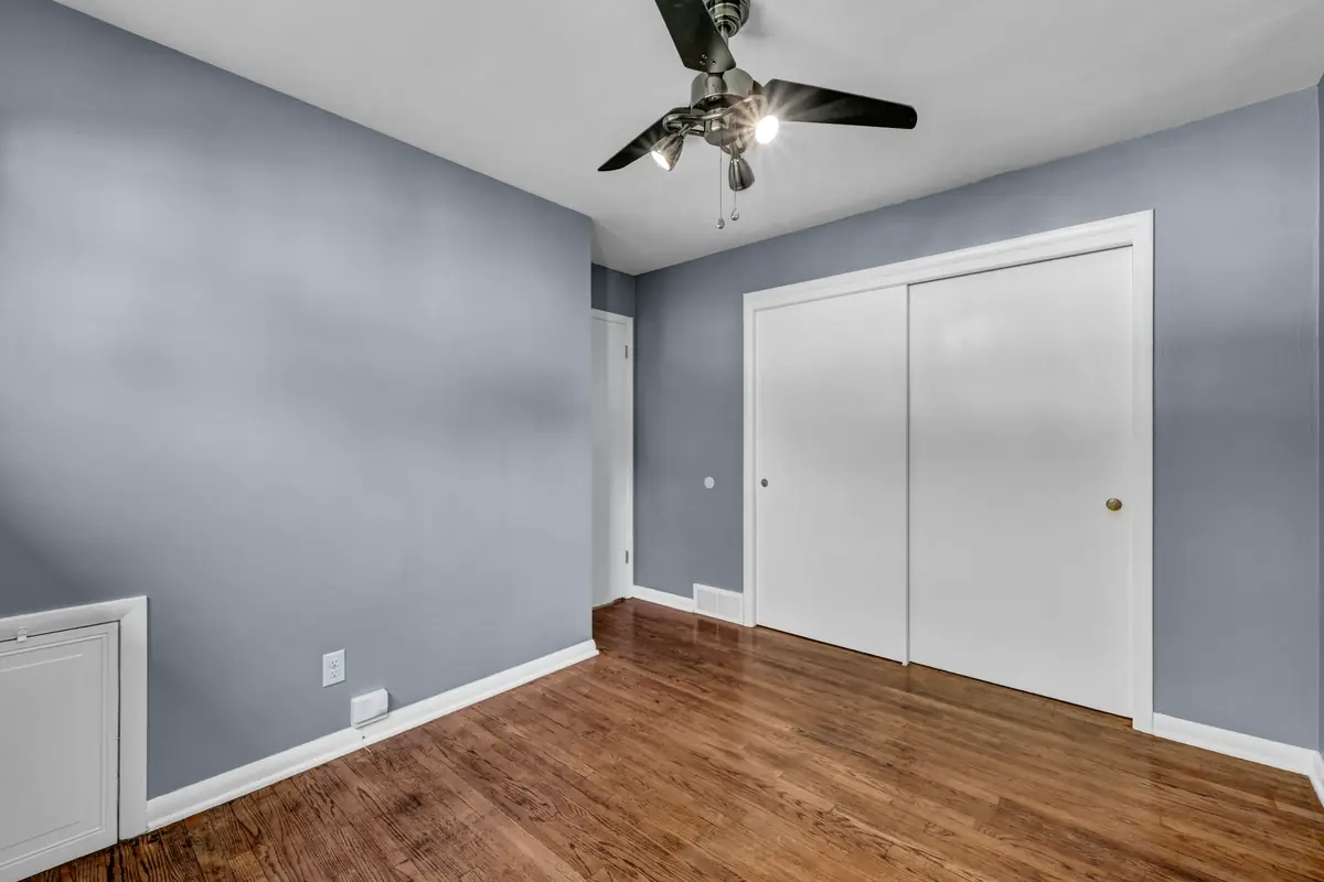 Empty bedroom with blue-gray walls, white sliding closet doors and baseboards, a ceiling fan and a wood floor