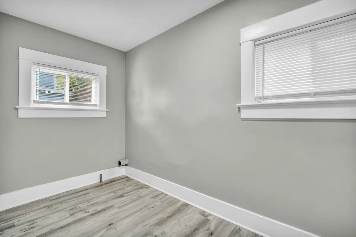 Corner of an empty room with light gray walls, two white-trimmed windows with blinds and gray plank flooring